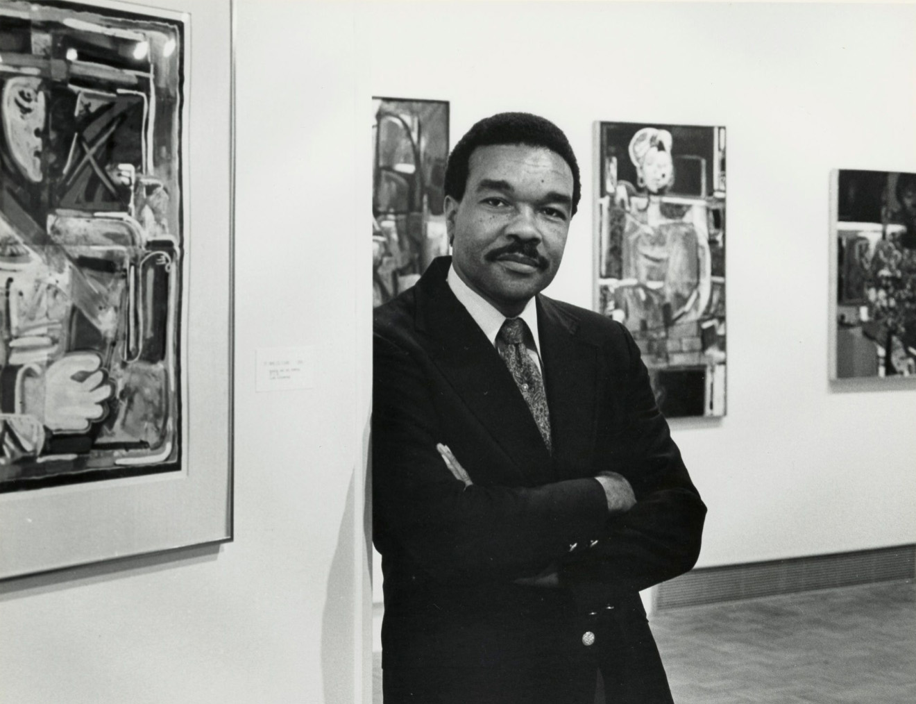 A man witha mustache in a suit leans with crossed arms against a wall in an art gallery