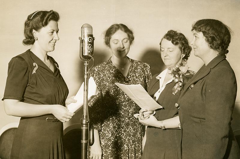Four women hold scripts and cluster around a microphone