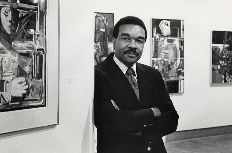 man in a suit with a mustache leans against a wall with his arms crossed in an art gallery
