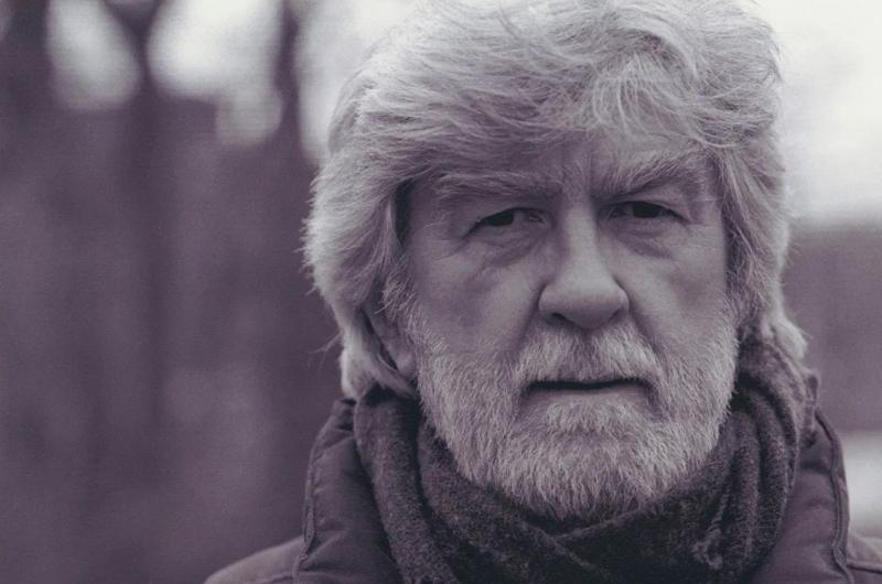 Stanley Plumly, an older man with a white beard and hair, looks into the camera with a serious expression, wearing a scarf